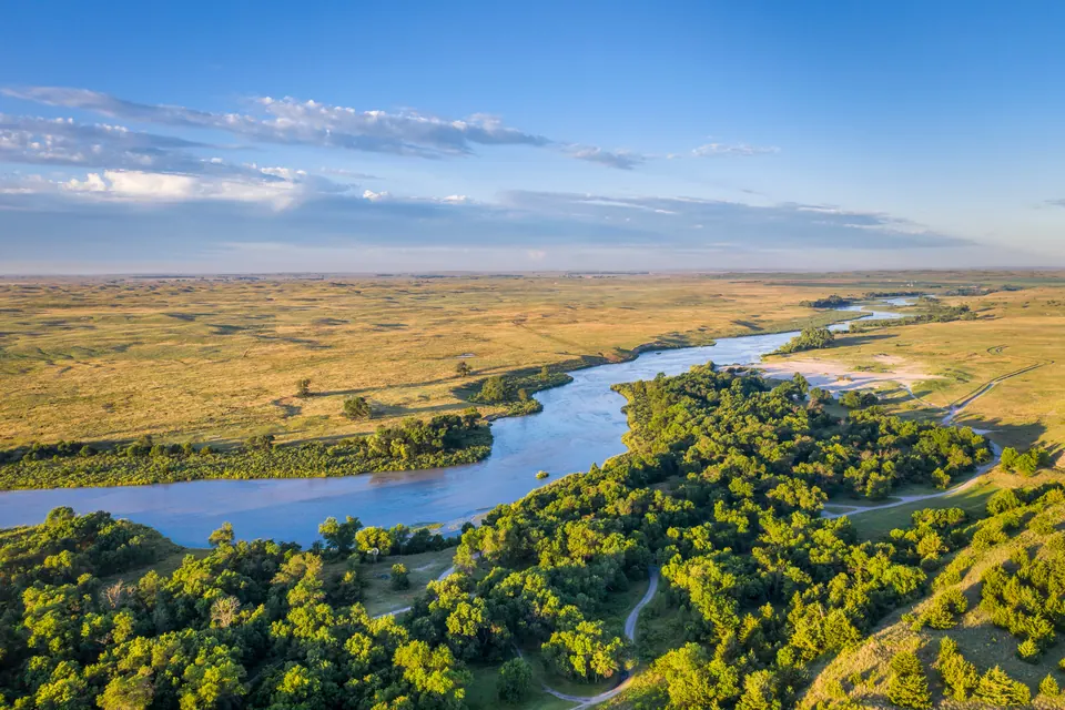 Luftaufnahme eines breiten, flachen Flusses; im Vordergrund Laubbäume und ein Feldweg, am anderen Ufer eine helle Sandbank, dahinter weites, grasbewachsenes Hügelland.