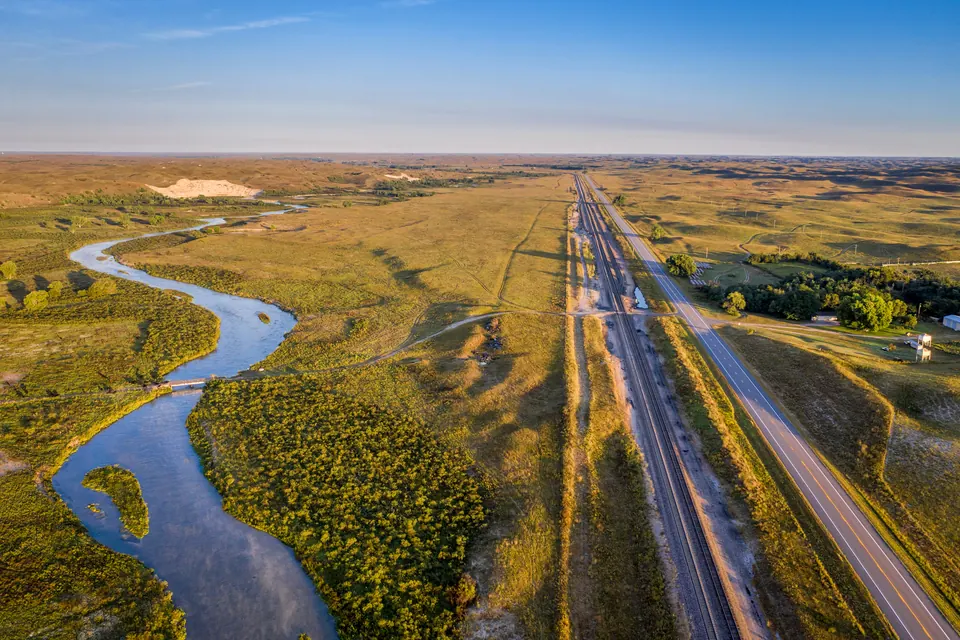 Luftaufnahme: links ein gewundener Fluss zwischen Buschwerk, in der Mitte eine Bahnstrecke, rechts eine Landstraße mit einem Gehöft, dahinter grasbewachsene Hügel bis zum Horizont; an einer Stelle liegt heller Sand offen.