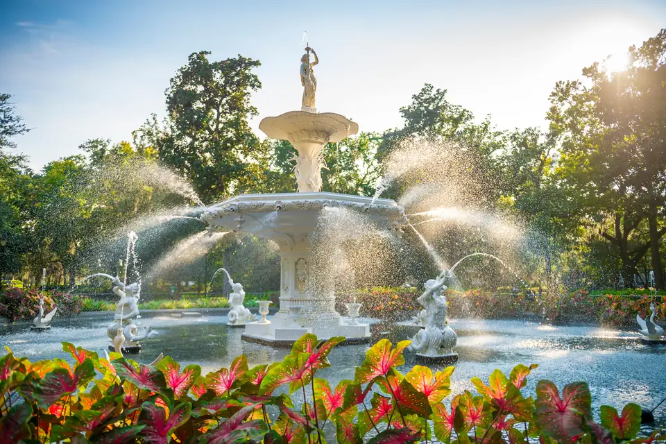 Ein weißer, mehrstufiger Brunnen mit einer Figur an der Spitze und wasserspeienden Figuren im Becken. Vorn wachsen Pflanzen mit rot-grünen Blättern, dahinter stehen Bäume im Gegenlicht.