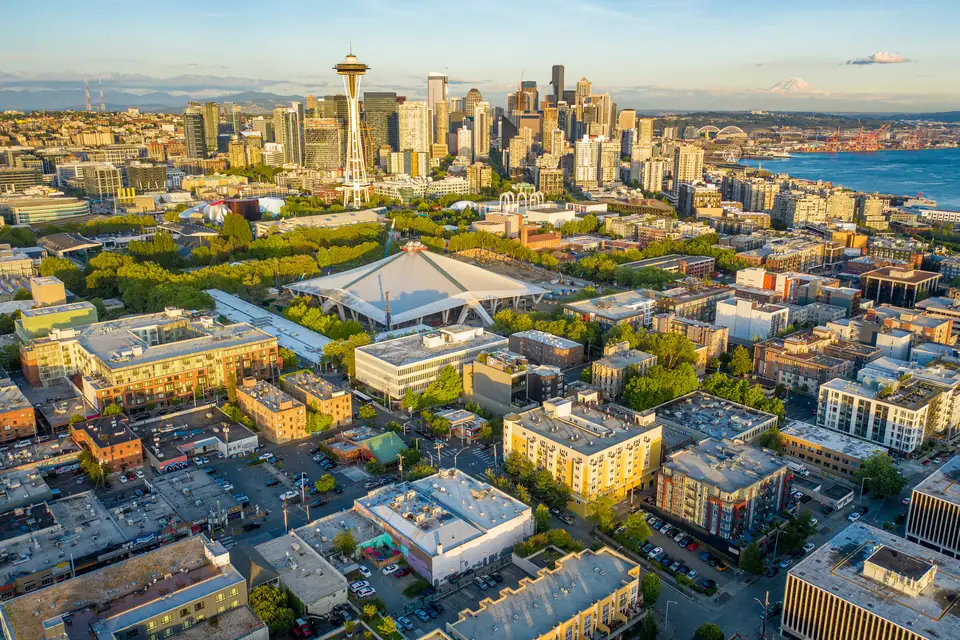 Luftbild bei tief stehender Sonne. In der Mitte liegt ein großes, spitz zulaufendes Hallendach zwischen Bäumen. Dahinter stehen die Space Needle, ein Gebäude mit weißen Spitzbögen und die Hochhäuser der Innenstadt.