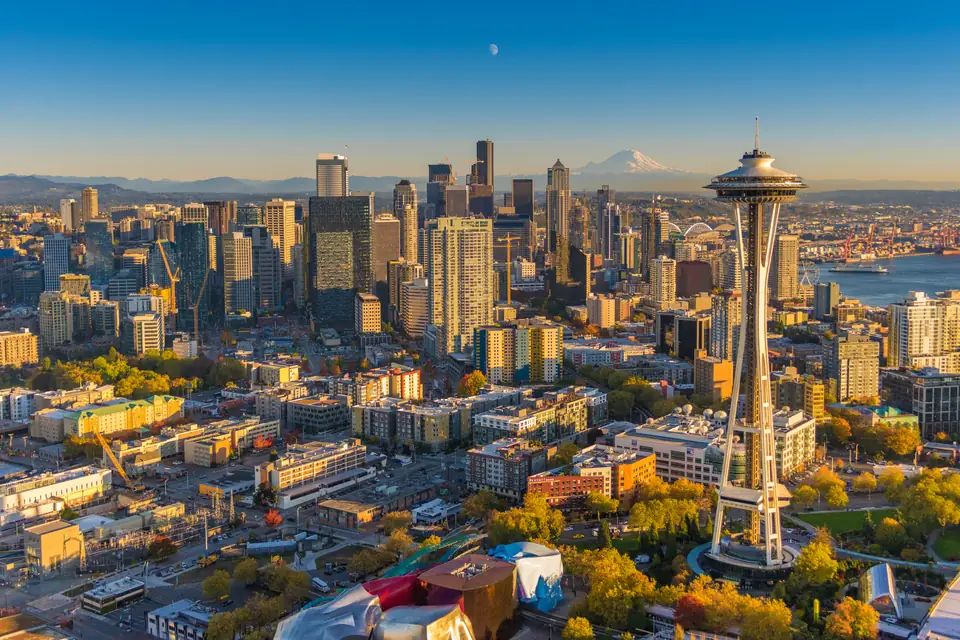 Luftbild im warmen Abendlicht: die Space Needle vor den Hochhäusern der Innenstadt, am Horizont ein schneebedeckter Berg. Unten links steht ein Gebäude mit farbigen, gewellten Metallfassaden, rechts unten ein Glasbau.
