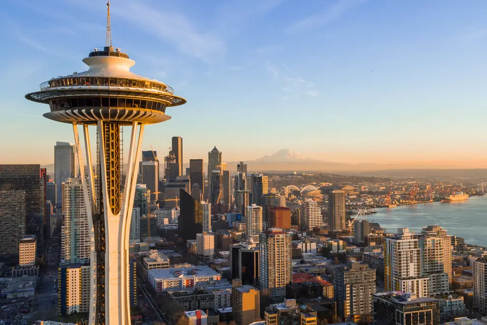Der Kopf der Space Needle mit umlaufender Aussichtsplattform in Nahaufnahme. Dahinter liegen Hochhäuser, eine Bucht und in der Ferne ein schneebedeckter Berg im Dunst.