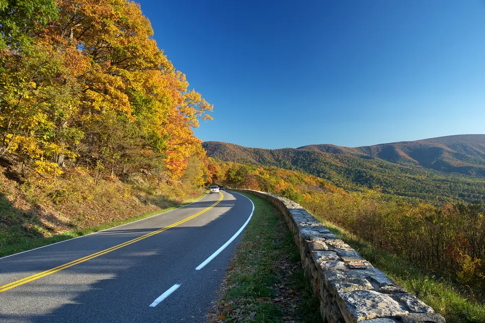 Eine zweispurige Straße mit gelber Mittellinie und Steinmauer führt an herbstlich gefärbtem Wald vorbei, dahinter Bergketten.
