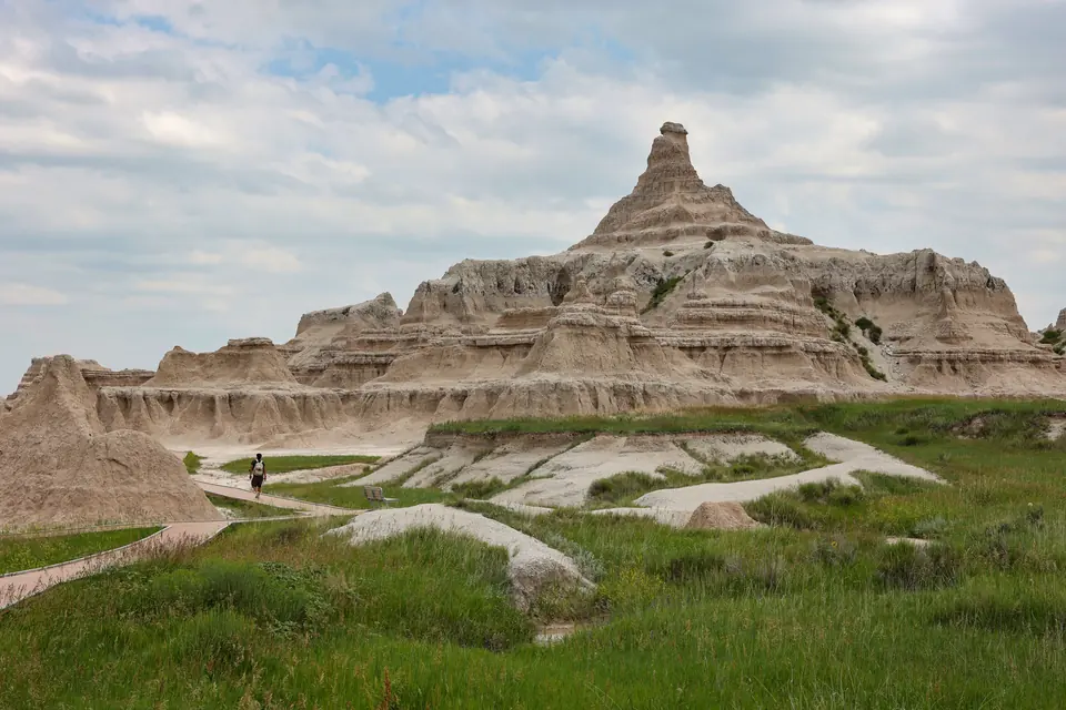 Gestufte Erosionsformationen mit einer spitzen Kuppe, davor Grasland mit einem schmalen Weg, auf dem eine Person geht.