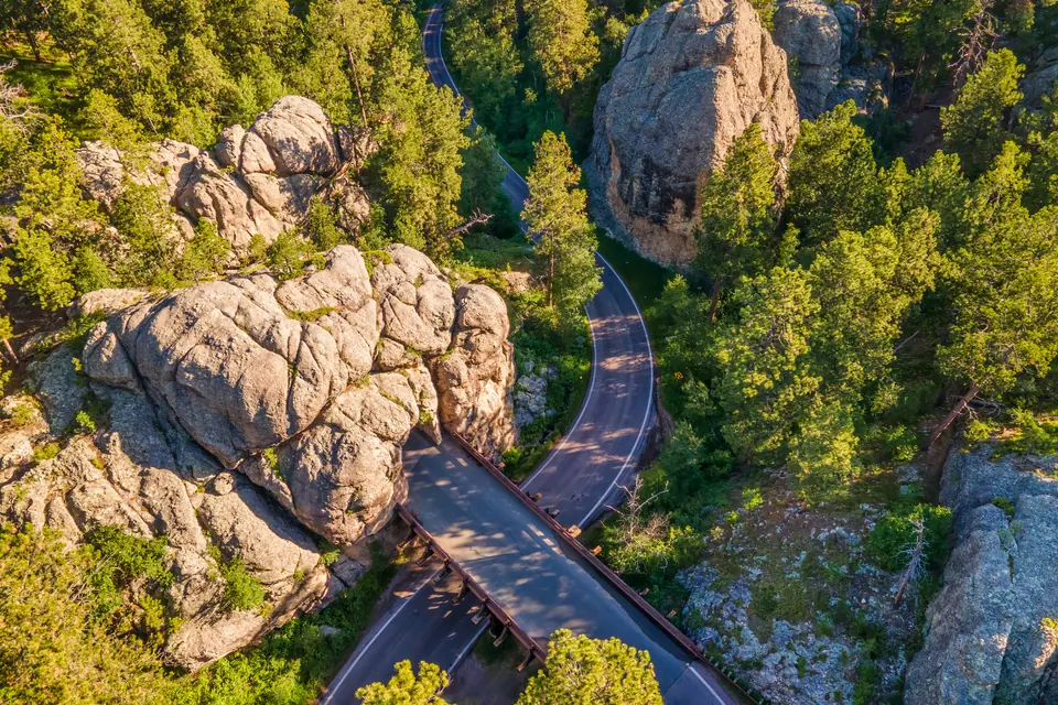 Luftbild einer Straße, die sich zwischen Granitfelsen und Kiefernwald windet, im Vordergrund eine Holzbrücke in einer Schleife.