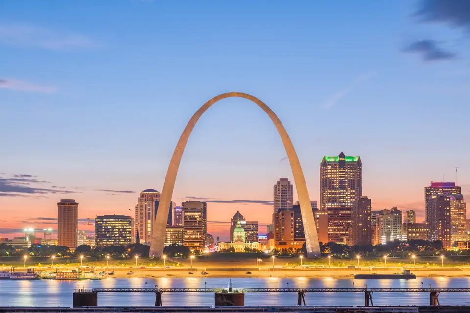Der Gateway Arch und die beleuchtete Skyline von St. Louis in der Abenddämmerung, gesehen über den Mississippi, auf dem sich die Lichter spiegeln.