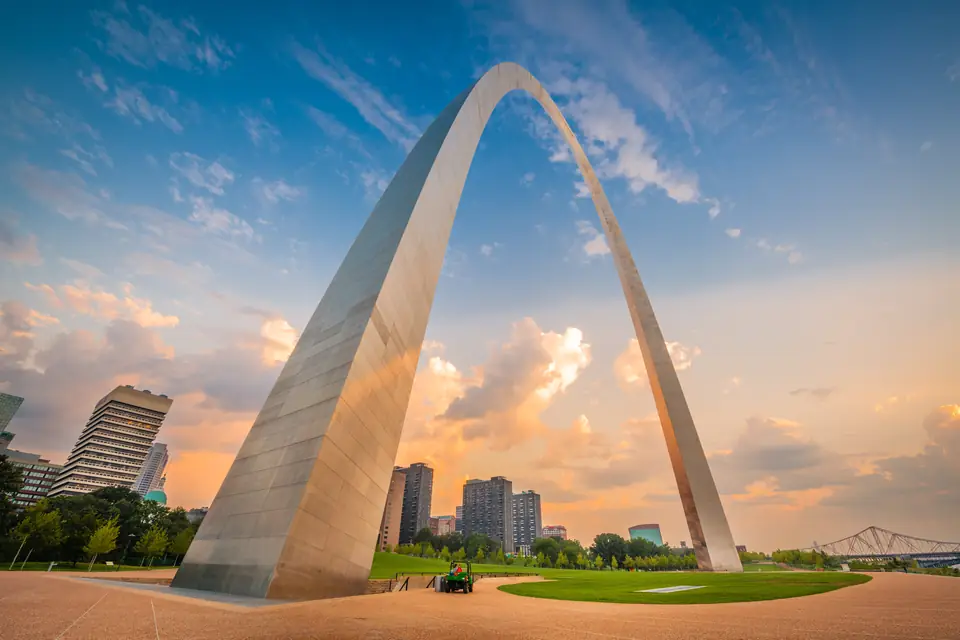 Ein Schenkel des silbernen Gateway Arch ragt schräg in einen Himmel mit rosa Wolken; dahinter Hochhäuser und eine Brücke.
