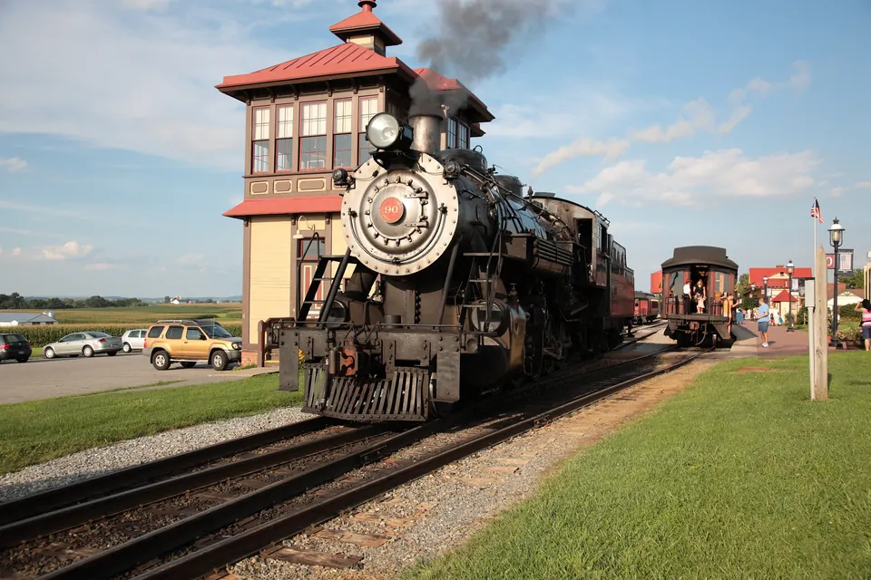 Eine schwarze Dampflokomotive mit der Nummer 90 an der Rauchkammertür steht mit Personenwagen an einem Bahnsteig, dahinter ein Stellwerkturm mit rotem Dach, ein Parkplatz und Maisfelder.