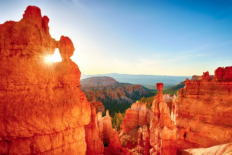 Orangerote Felstürme im Gegenlicht der tief stehenden Sonne, dahinter ein bewaldetes Tal.