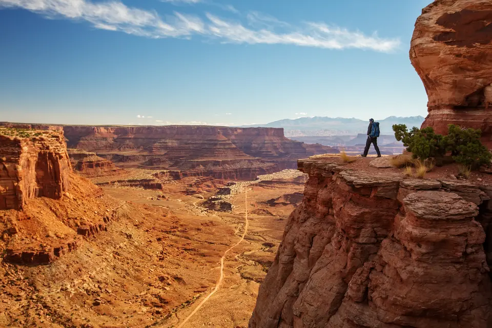 Eine Person steht an der Kante einer Felsplattform über einem weiten Canyon, unten zieht sich eine Piste durch das Tal.