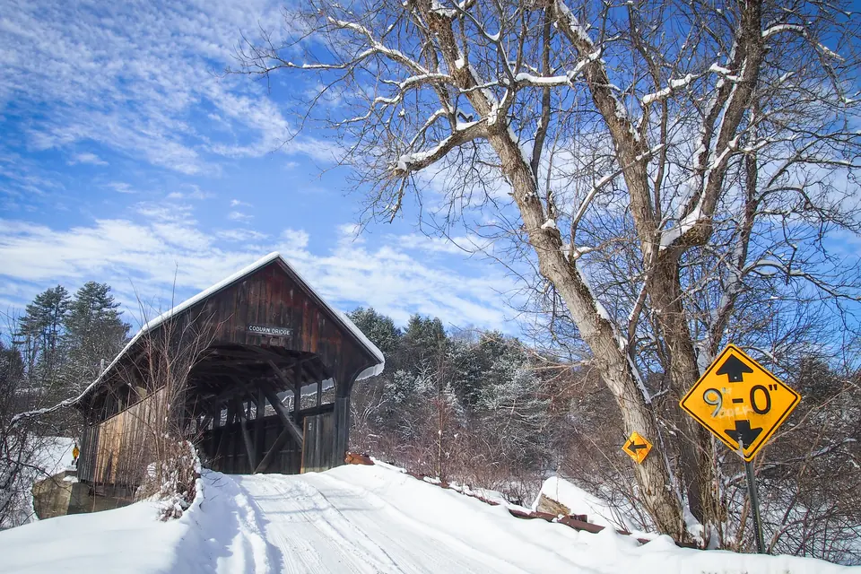 Eine gedeckte Holzbrücke mit verschneitem Dach am Ende einer schneebedeckten Straße, daneben kahle Laubbäume und Nadelbäume unter blauem Himmel, rechts ein gelbes Höhenschild.