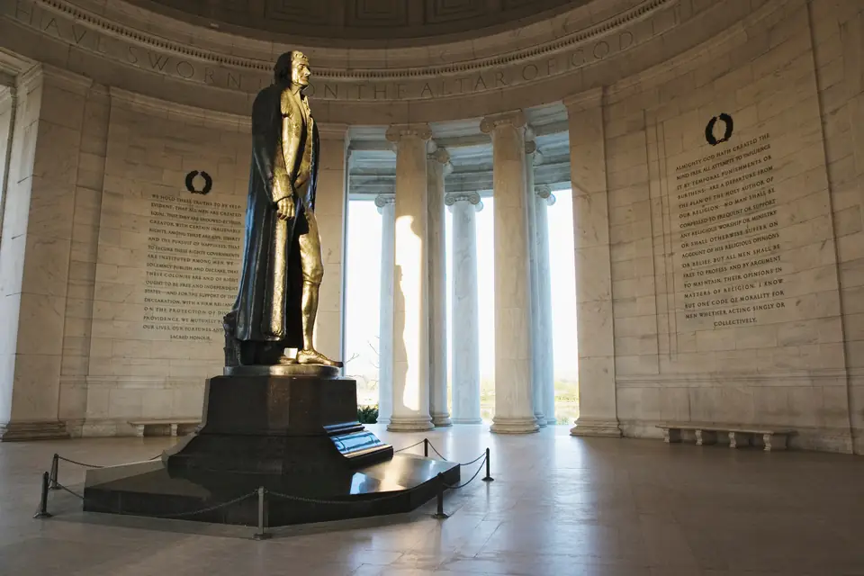 Innenraum des Jefferson Memorial: eine große dunkle Statue Thomas Jeffersons auf einem Sockel hinter einer Absperrkette, dahinter hohe Säulen mit Durchblick nach draußen und Wände mit eingemeißelten Texten.