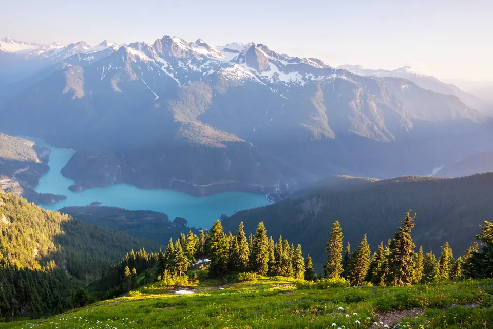 Türkisfarbener See zwischen bewaldeten Hängen, dahinter schneebedeckte Gipfel, im Vordergrund eine Bergwiese mit Nadelbäumen.