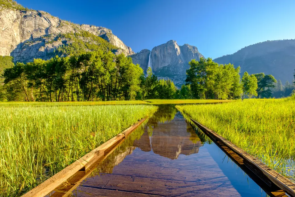 Ein hölzerner Bohlenweg steht in einer grünen Wiese unter Wasser, im Wasser spiegelt sich eine Felswand, dahinter fällt ein Wasserfall über die Granitwand.