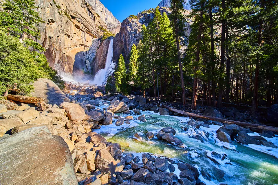 Ein Wasserfall stürzt zwischen hohen Granitwänden herab, davor ein Bach über große Steine, an seinen Rändern weißer Reif und Eis, rechts Nadelbäume.
